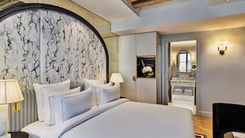 Una habitación de hotel moderna con cabecera de mármol, una cama grande, una zona de estar con una silla y un baño visible a través de una puerta abierta.