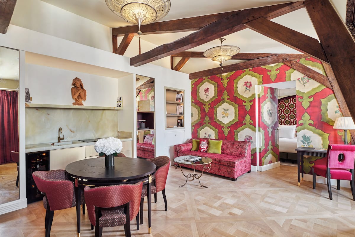 A spacious loft with exposed wooden beams, a dining area with a round table and chairs, a red patterned wall, and a kitchen with a marble countertop.