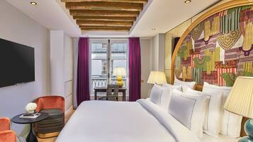 A hotel room with a large bed, a television, a small table with a lamp, and a view of the city through the window.