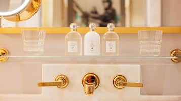 Un baño con un espejo de marco dorado, tres botellas de agua de colonia y tres vasos de vidrio transparente sobre una estantería.