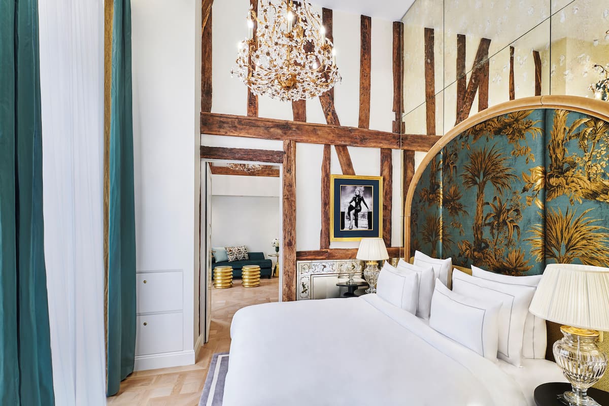 A bedroom with a large bed, a chandelier, a framed picture on the wall, and a view into another room.