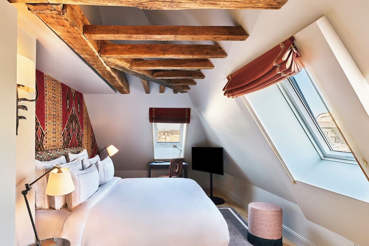 A bedroom with a large bed, a desk, a chair, a TV, a window with curtains, and a skylight.