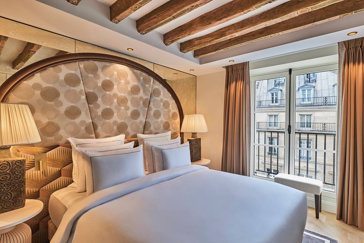 A hotel room with a large bed, two bedside tables with lamps, a round headboard, and a view of the city through large windows.