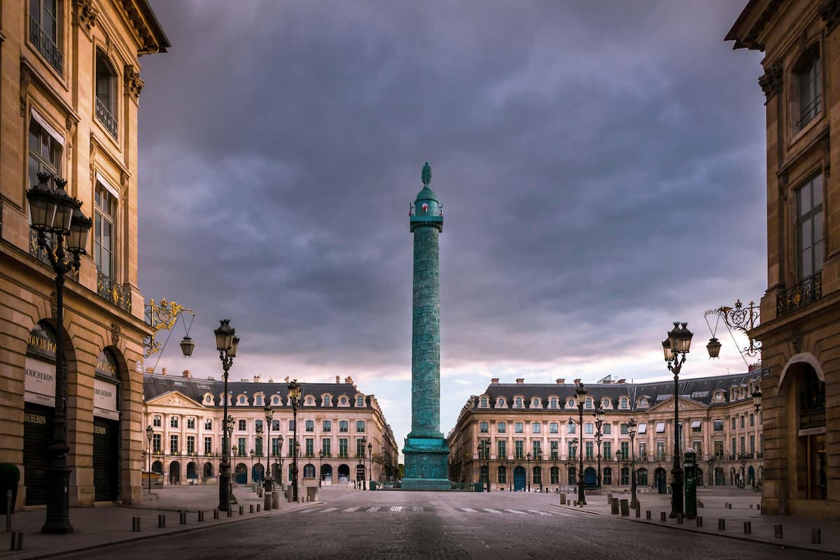 A historic square with a central monument, classical architecture, and a paved ground.