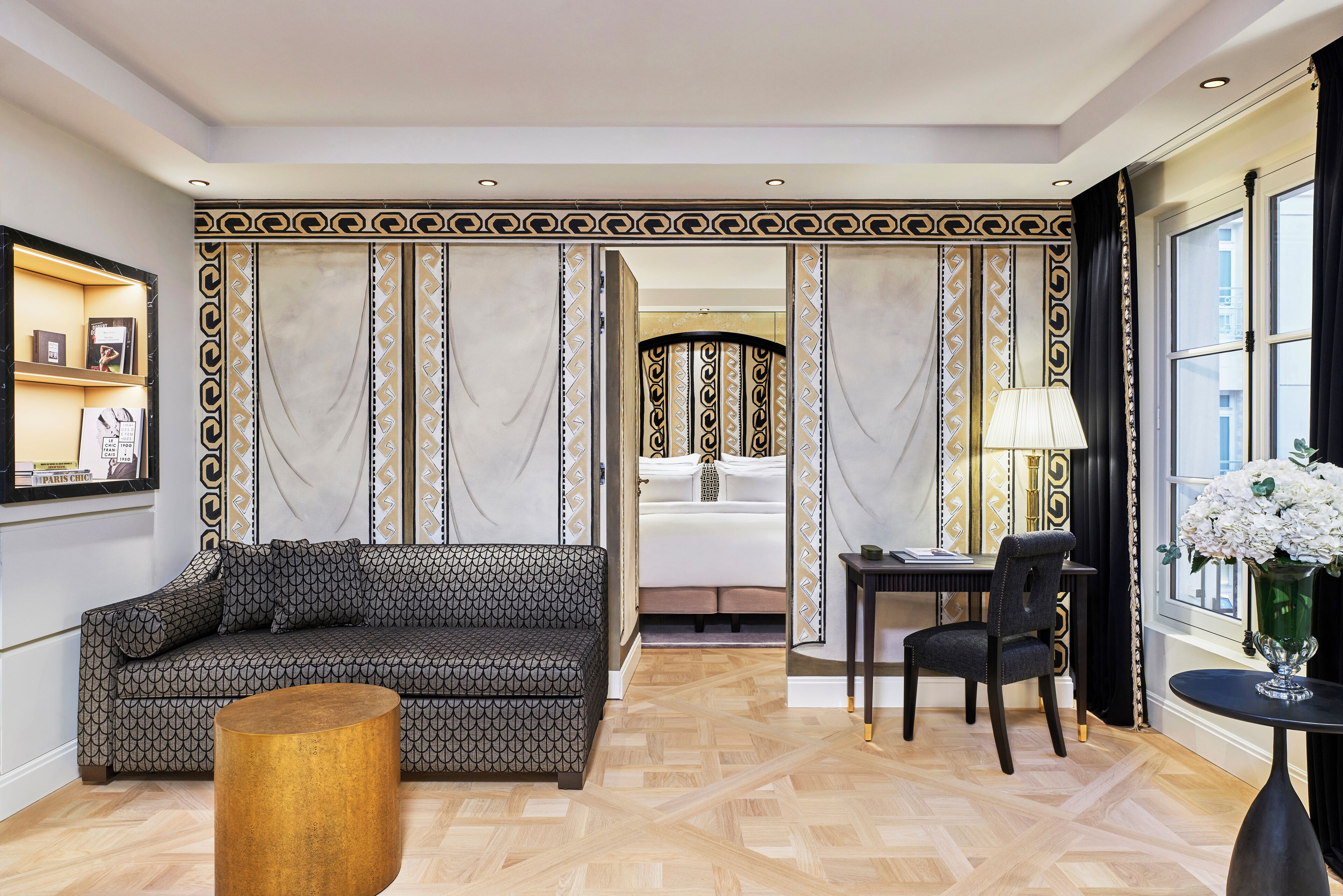A modern hotel room with a patterned sofa, a wooden coffee table, a dining area with a chair, and a bed with curtains.