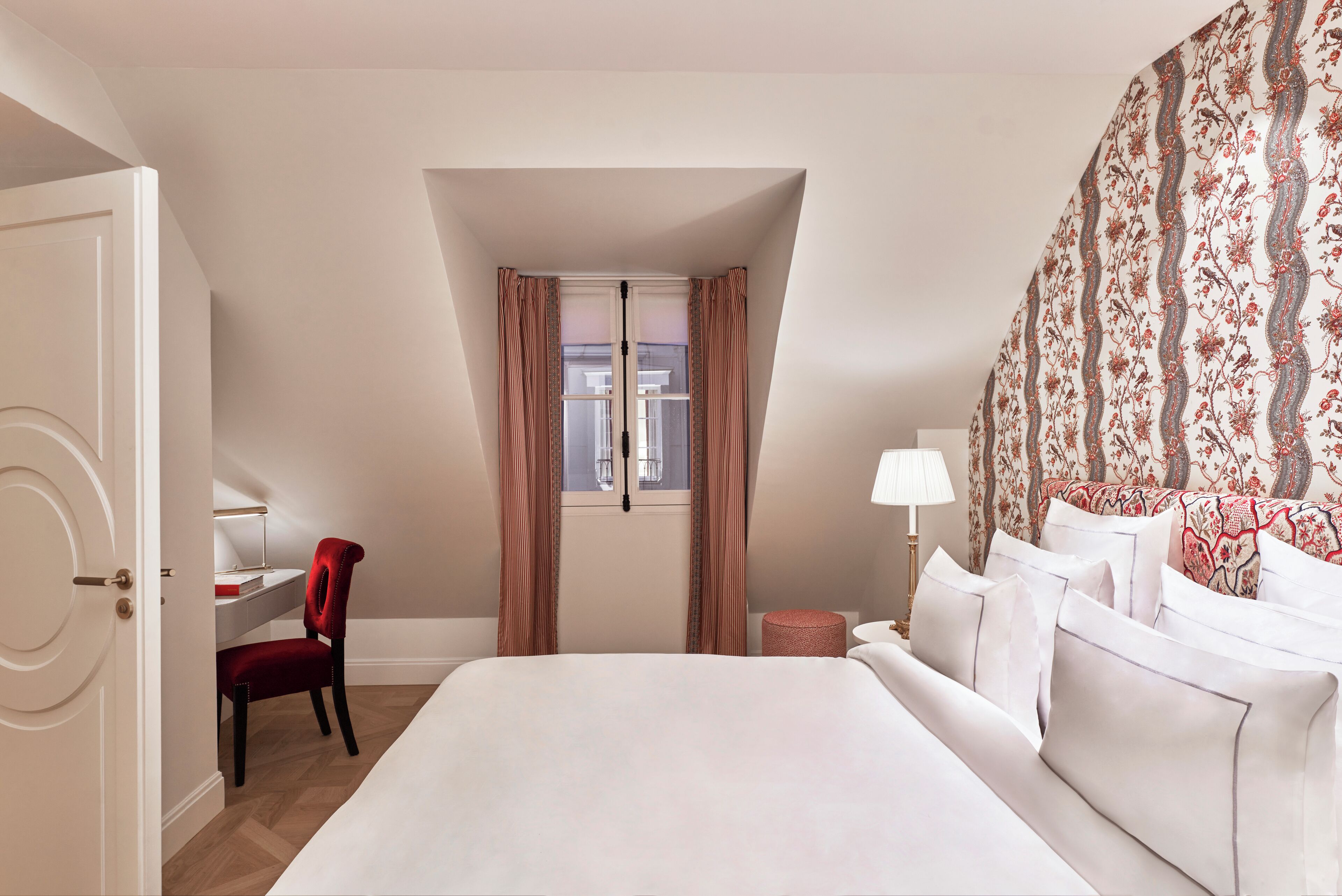 A hotel room with a large bed, a desk with a chair, a window with curtains, and a patterned wall.