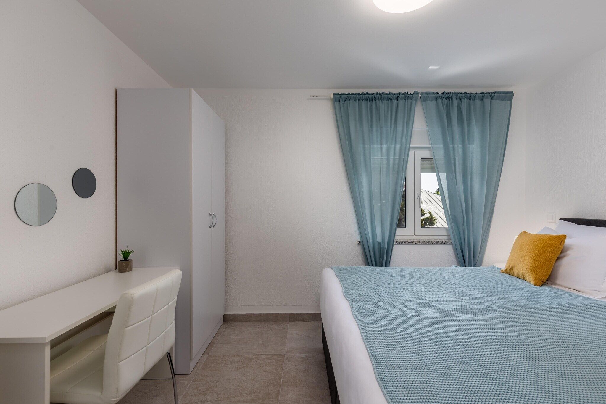 Un dormitorio con cama, escritorio, silla, planta pequeña y ventana con cortinas.