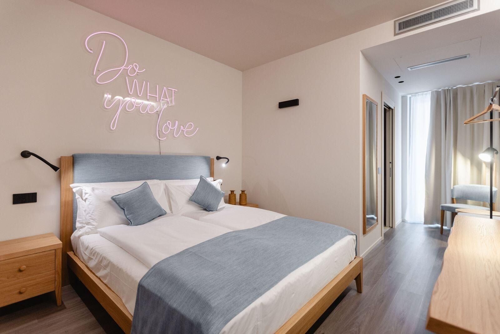 A modern bedroom with a bed, bedside tables, a bedside lamp, a neon sign, and a view of a hallway and a window.