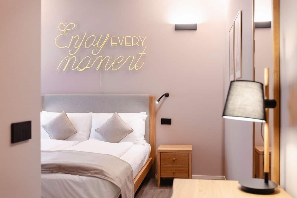 A neatly arranged bedroom with a bed, bedside table, lamp, and a neon sign on the wall.