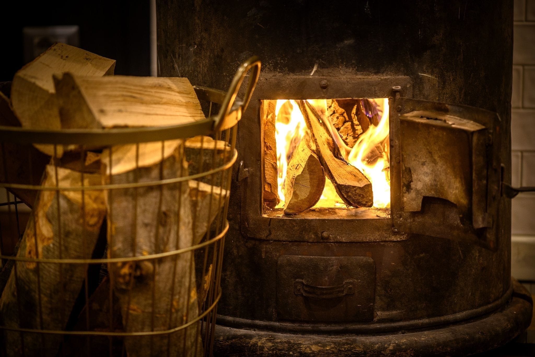 A wood-burning stove with logs burning inside, a metal basket with logs outside, and a metal handle on the stove.