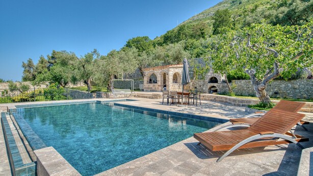 Une piscine avec des chaises longues, un bâtiment en pierre doté d’arcades et une colline boisée.