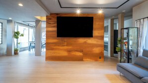 Un salón amplio con una televisión de pantalla plana grande montada en una pared de madera, un sofá gris y una partición de cristal con plantas.