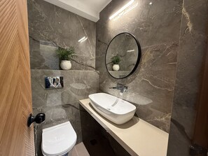 Ein modernes Badezimmer mit einer Marmorwand, einem runden Spiegel, einem weißen Waschbecken und einer Toilette.