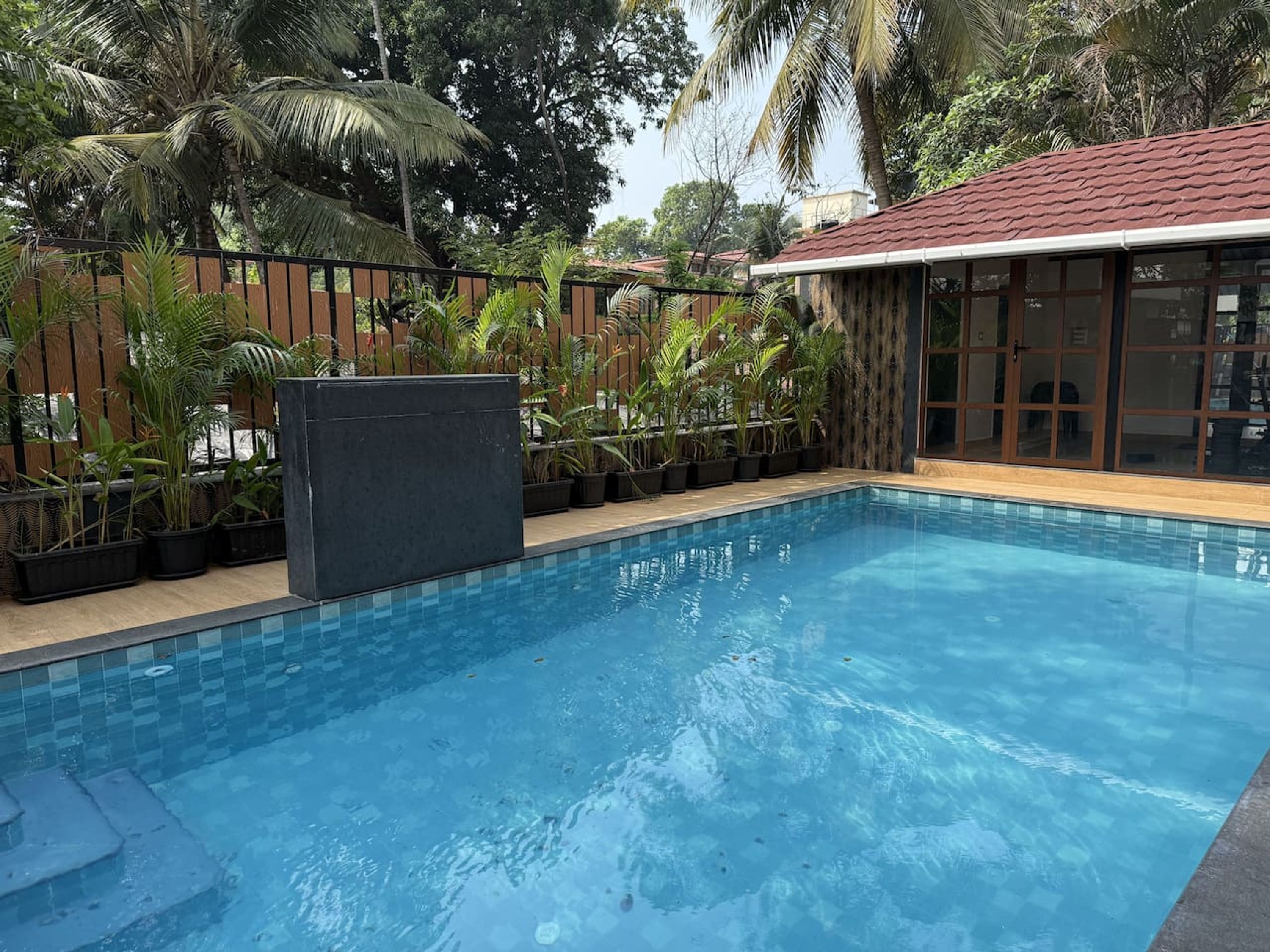 A swimming pool with a tiled deck, surrounded by a wooden fence and various tropical plants.