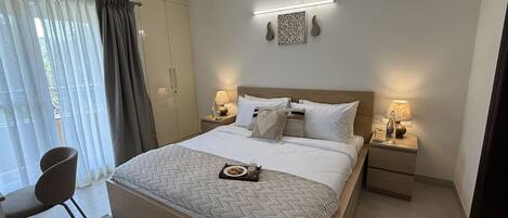 A modern hotel room with a large bed, two bedside tables with lamps, a wardrobe, and a seating area with a chair and a small table.