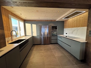 A modern kitchen with a stainless steel refrigerator, oven, and sink, wooden ceiling, and large windows.