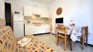A compact living space with a sofa, a dining table with chairs, a kitchen area with cabinets and appliances, and a decorative wall piece. - Adorable apartment with outdoor pool (Bibione)
