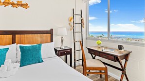 A hotel room with a bed, a desk, a chair, a window with a view of the ocean, and a decorative wall piece. - Ocean View 1BR at Ilikai Marina  (Honolulu)