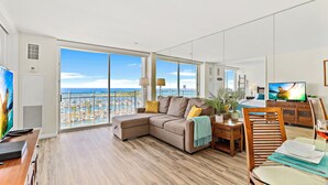 A spacious living room with a large sectional sofa, a dining table, and a TV. The room has large windows offering a view of the sea. - Ocean View 1BR at Ilikai Marina  (Honolulu)