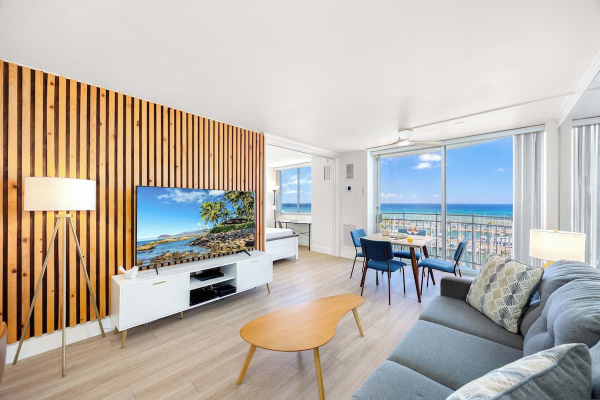 Ein modernes Wohnzimmer mit großem Fernseher, einer Wand aus Holzpaneelen und einem Balkon mit Blick auf den Strand.