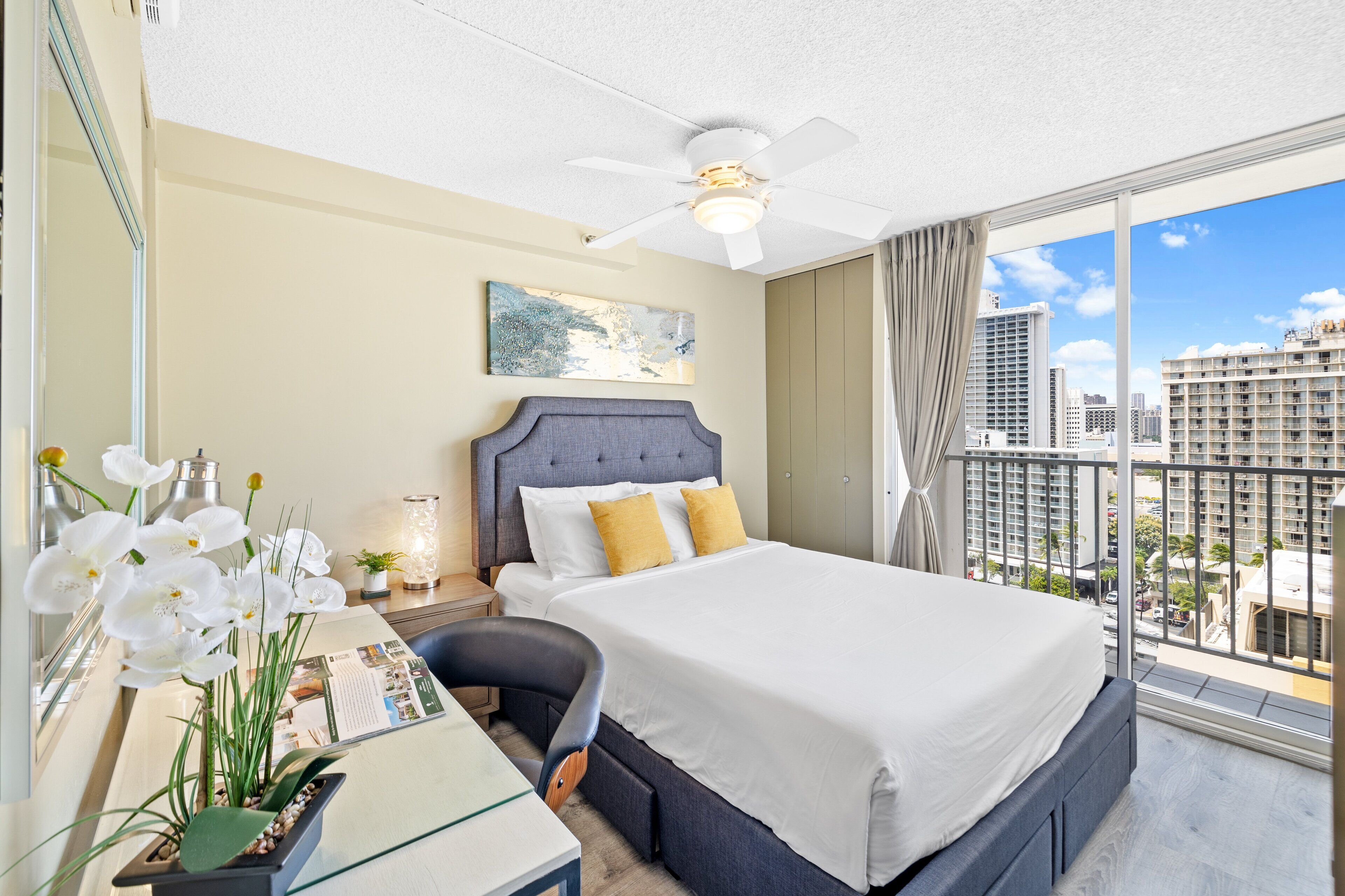 A hotel room with a large bed, a desk with a computer, a potted plant, and a city view through large windows.