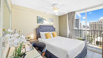 A hotel room with a large bed, a desk with a computer, a potted plant, and a city view through large windows.