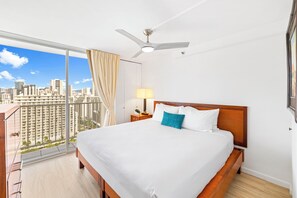 A hotel room with a large bed, a ceiling fan, a nightstand with a lamp, and a view of a cityscape through a large window. -  Pacific Monarch - 1 BR - Ocean & City Views (Honolulu)