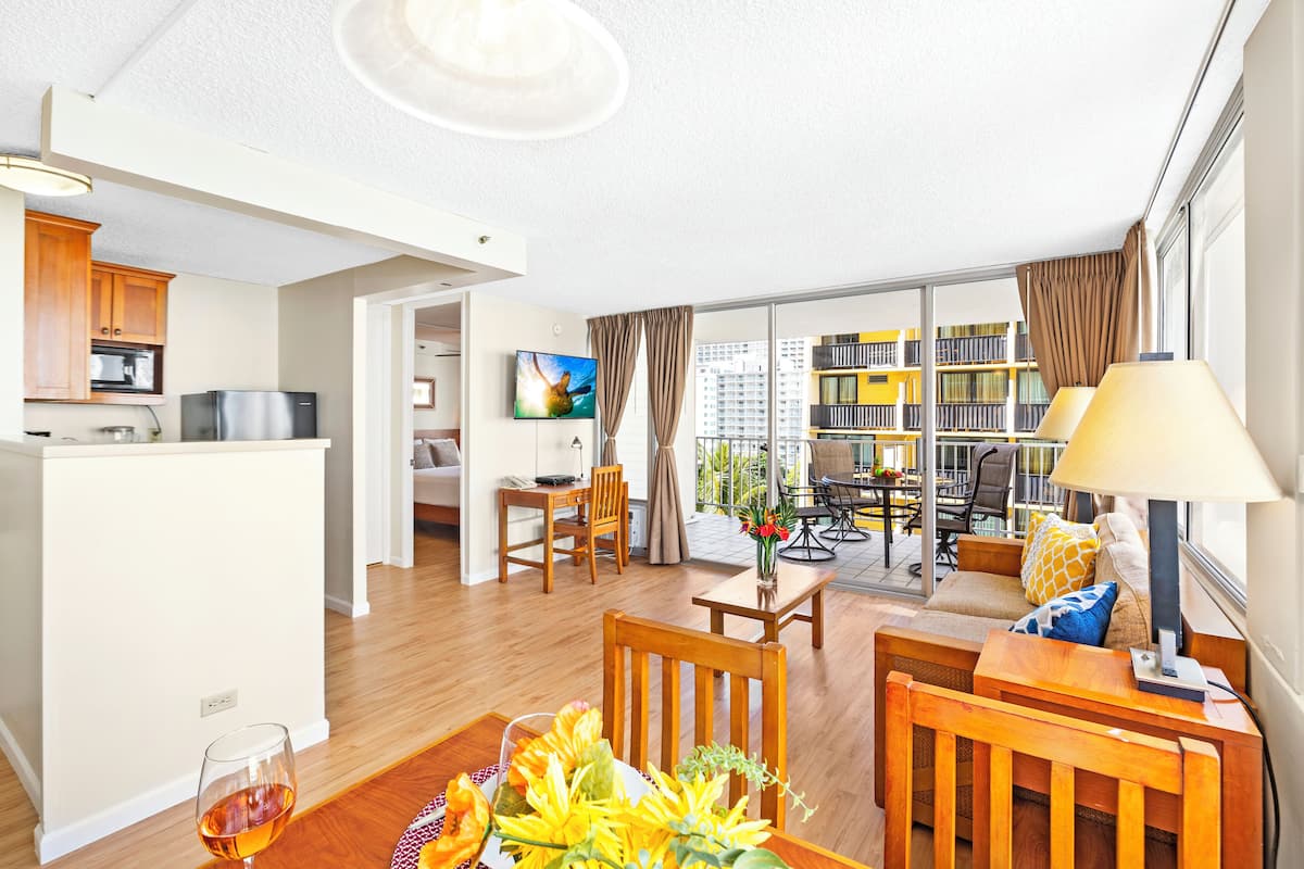 A modern hotel room with a kitchen, dining area, and a balcony with a city view.