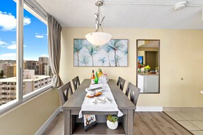 A dining area with a view of the city, a table set with dinnerware, and a painting of palm trees on the wall. -  Pacific Monarch - 1 BR - Ocean & City Views (Honolulu)