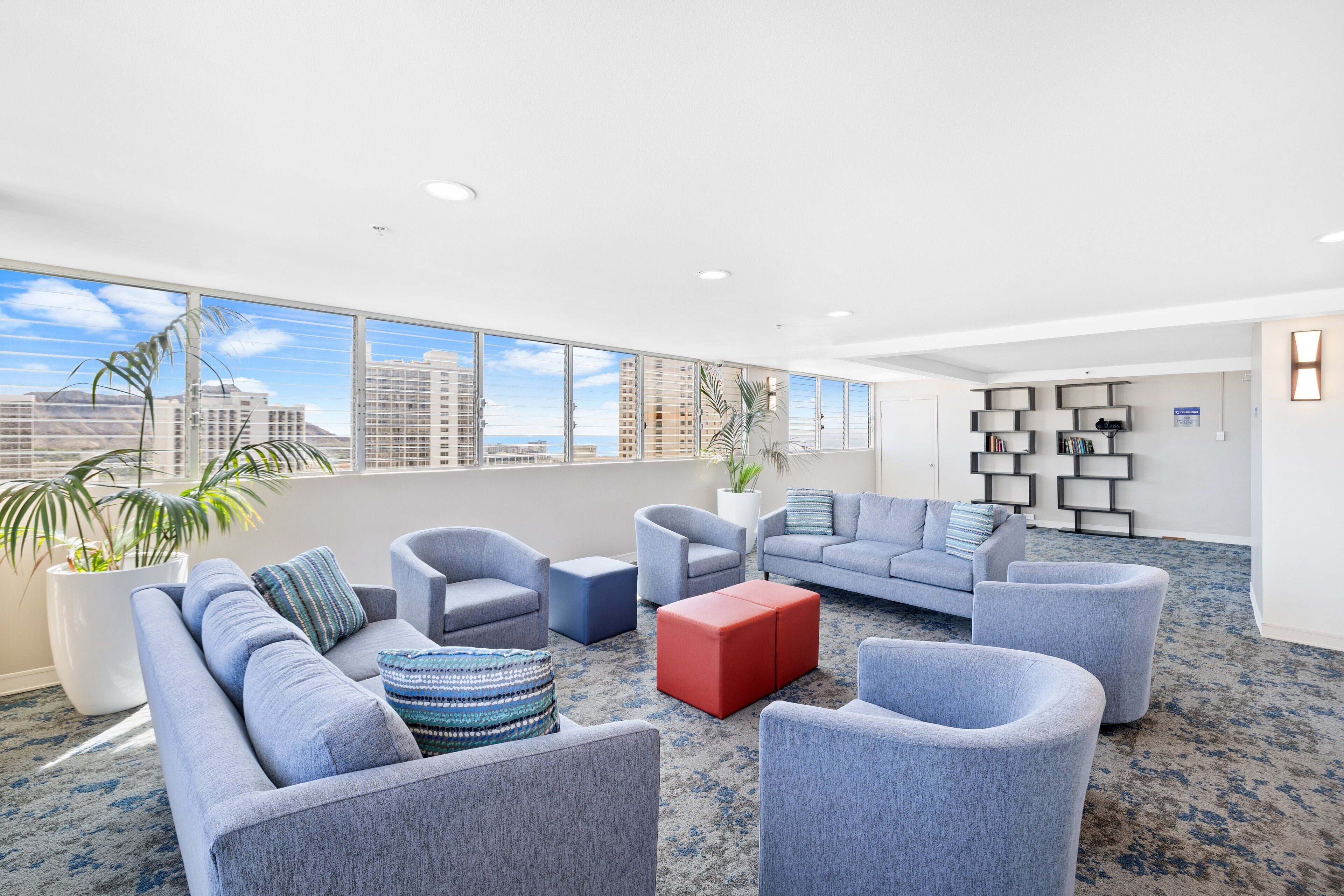 Ein stilvoller Lounge-Bereich mit grauen Sofas, Sesseln und Poufs, großen Fenstern mit Blick auf die Stadt und einem gemusterten Teppich.