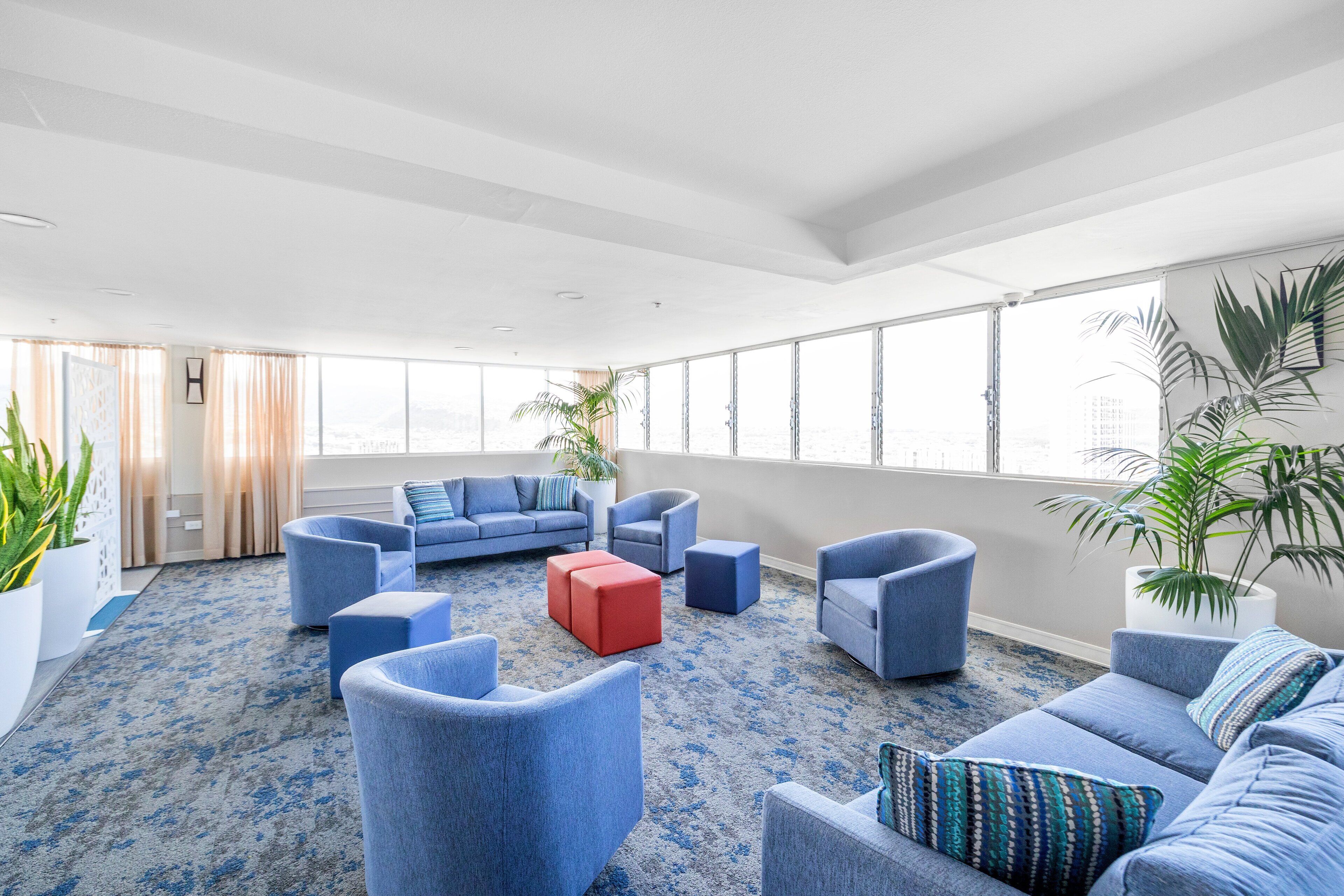 Een ruime lounge met blauwe banken en fauteuils, een rode poef en grote ramen met uitzicht op de stad.