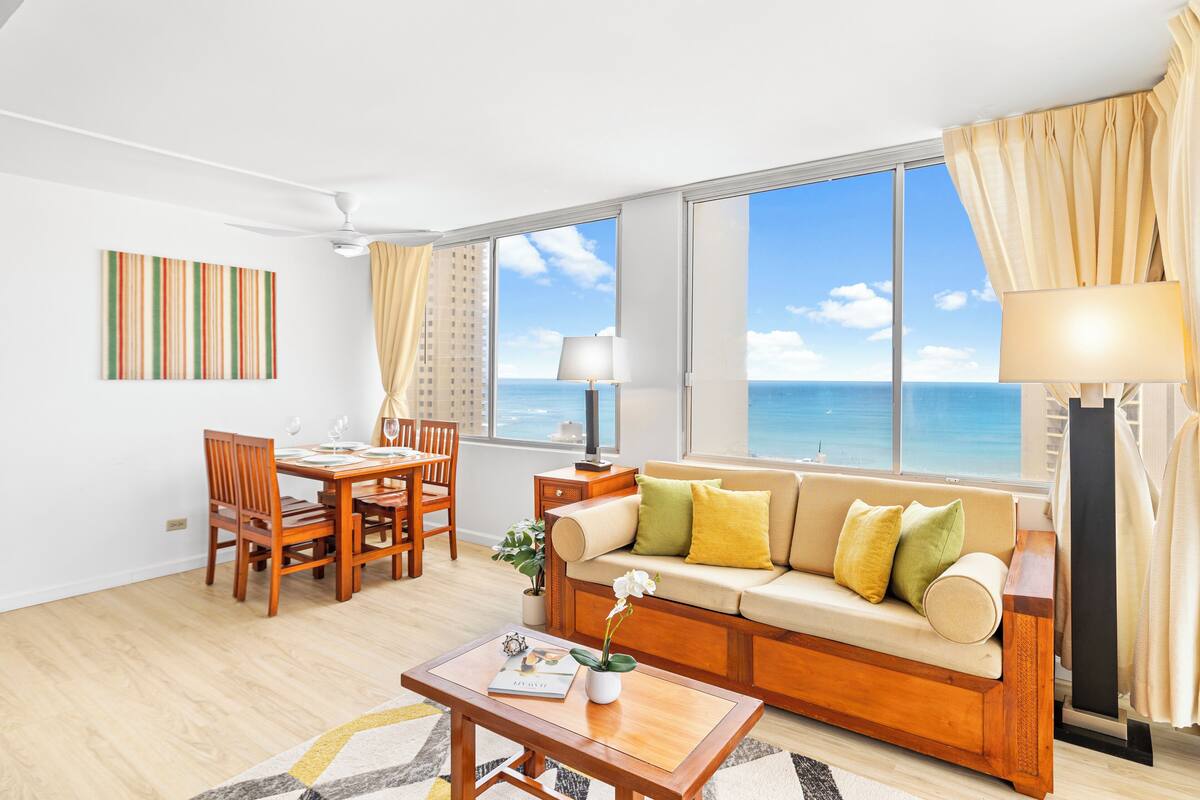 A modern living room with a sofa, dining table, and chairs. There is a large window with a view of the ocean.