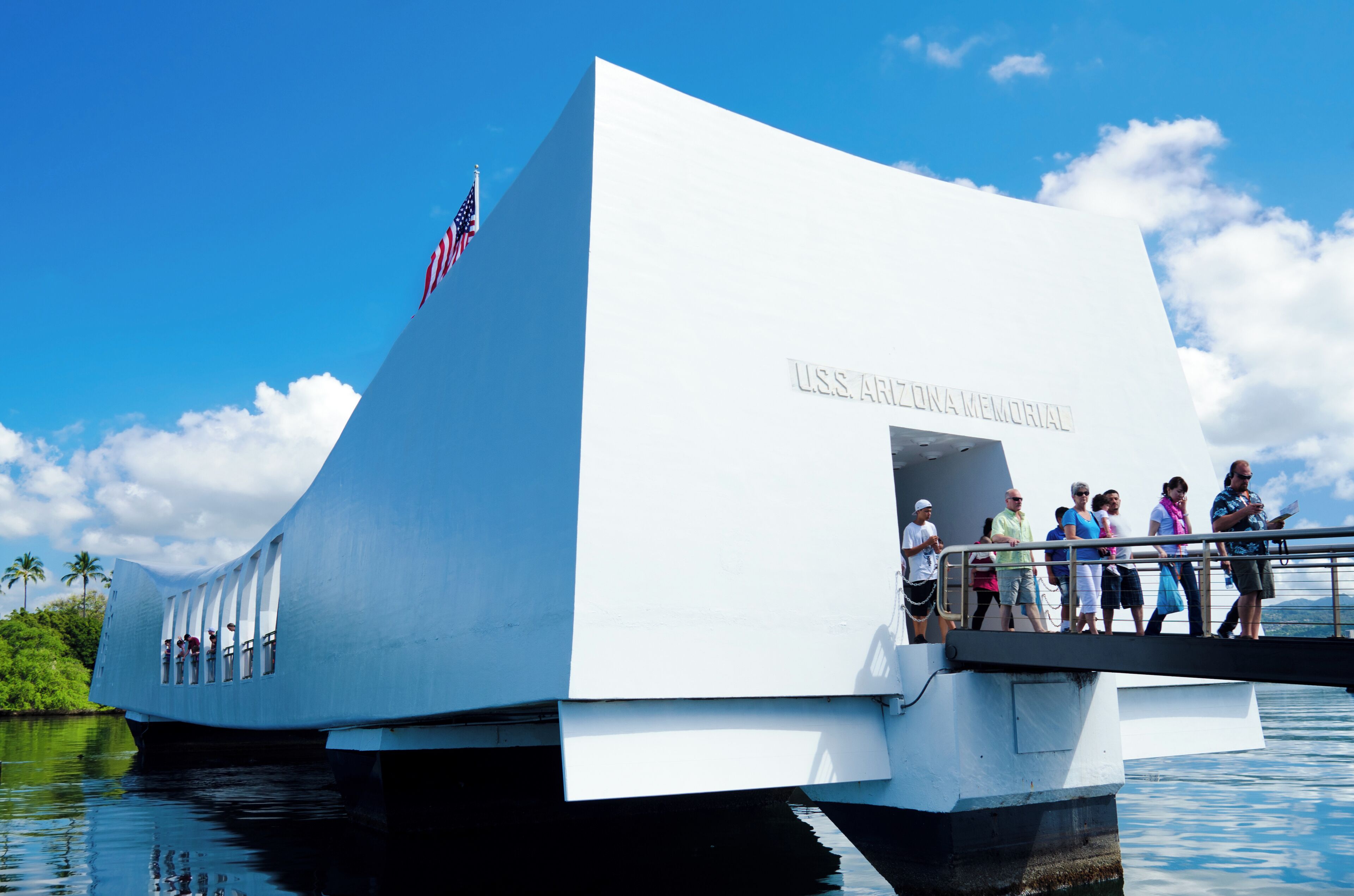 A large white building with "U.S.S. Arizona Memorial" written on it, located near water with people walking around.