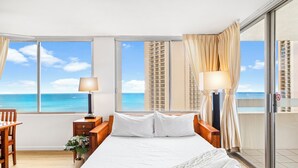 A hotel room with a large bed, two bedside tables, a desk, and a chair. There are large windows with curtains, a view of the ocean, and a balcony with a glass door.