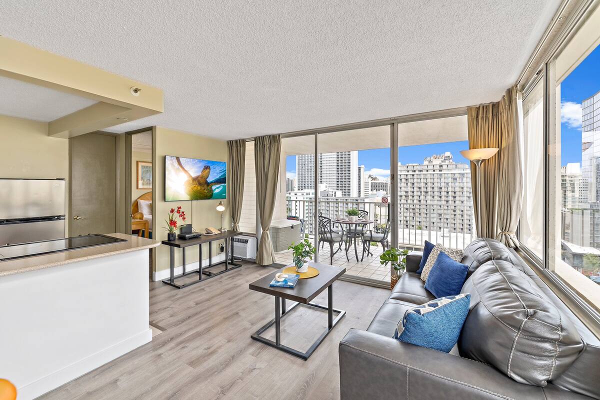 A modern apartment with a kitchen, dining area, and living room. It features a large window with a city view, a sofa with cushions, and a flat-screen TV.