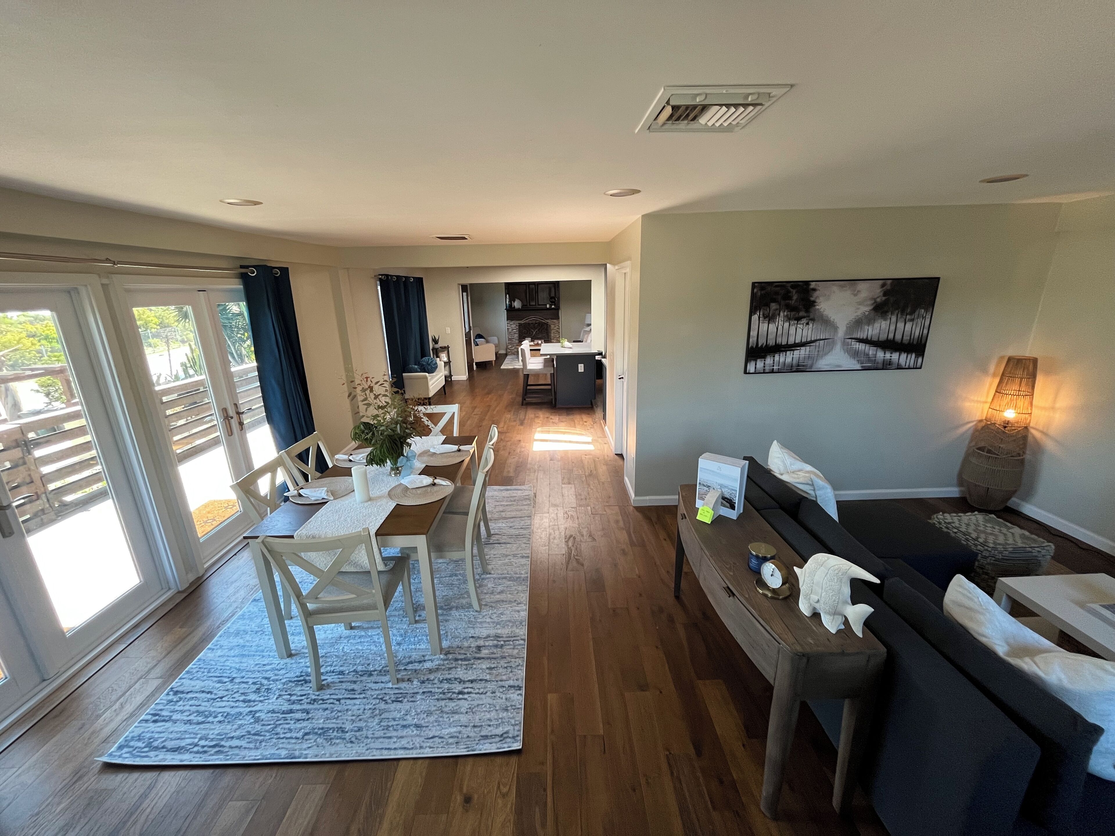 Un vaste espace de vie comprenant une table à manger pour quatre personnes, un canapé et une table basse. Au fond, on aperçoit une cuisine, une grande fenêtre avec des rideaux et un tableau accroché au mur.