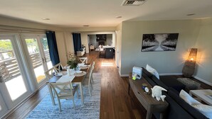 Een ruime woonkamer met een eettafel voor vier personen, een bank en een bijzettafeltje. Achtergrond is een keuken, een groot raam met gordijnen en een schilderij aan de muur.