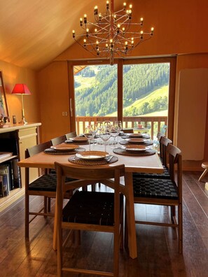 A dining area with a table set for four, a chandelier, and a view of a forested landscape through large windows.