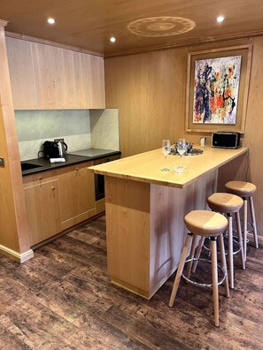 A modern kitchen with wooden cabinets, a central island with stools, a coffee maker, and a painting on the wall.