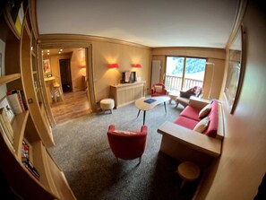 A cozy living room with a red armchair, a sofa, a small table, and a TV. There is a wooden cabinet and a door leading to another room.