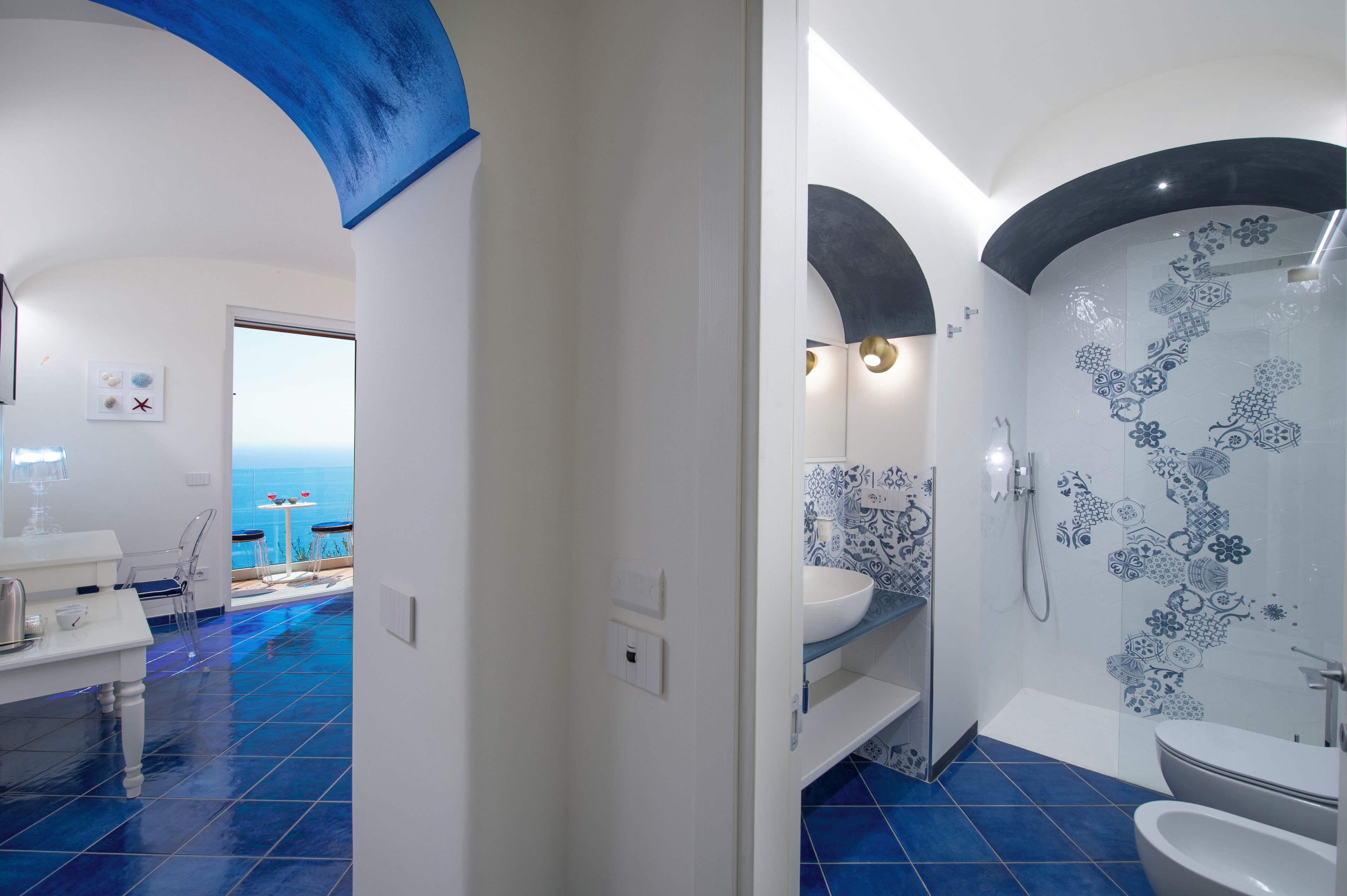 Un baño moderno con suelo de baldosas azules, un lavabo blanco, una zona de ducha con azulejos estampados y vistas al mar a través de una puerta abierta.
