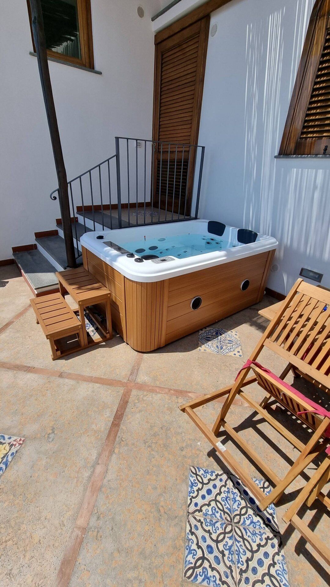 A hot tub with wooden steps and a metal railing, surrounded by wooden furniture and a tiled floor.