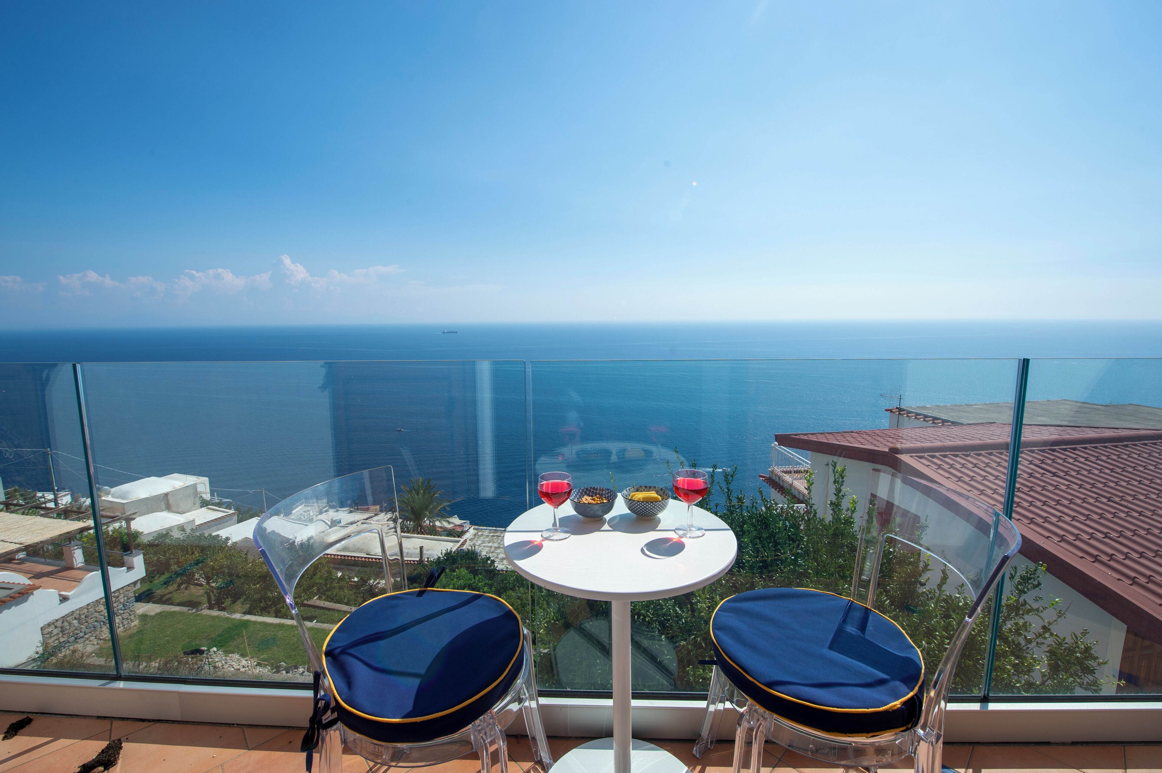 Un balcón con barandilla de vidrio que ofrece vistas al mar. Hay una mesa pequeña con dos sillas y un cuenco de fruta.