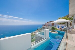 Una piscina infinita en la azotea con vistas al océano, un pequeño jardín y mobiliario de exterior.