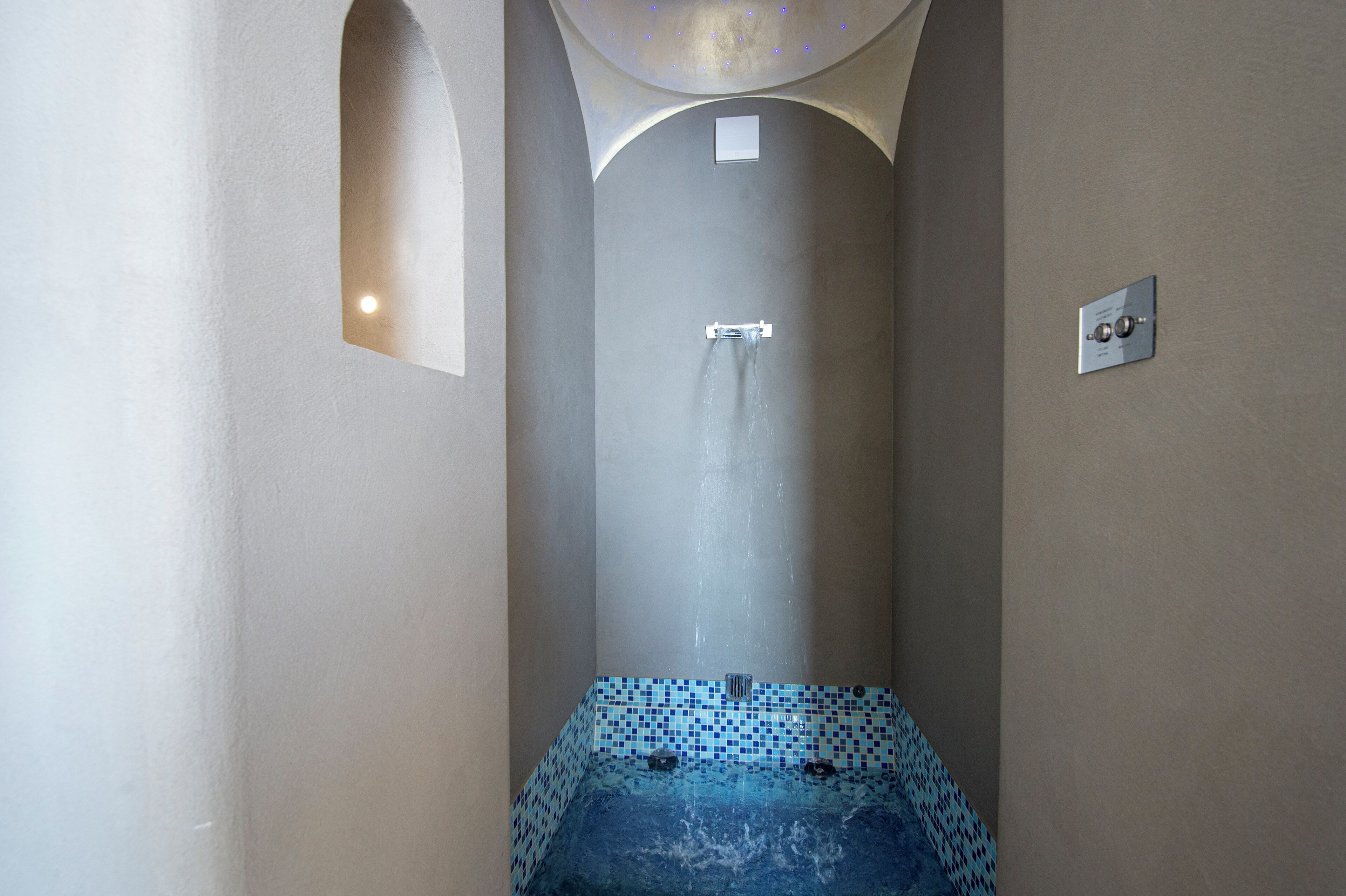 Un espacio de ducha estrecho con azulejos, una única salida de agua y un panel de control en la pared.