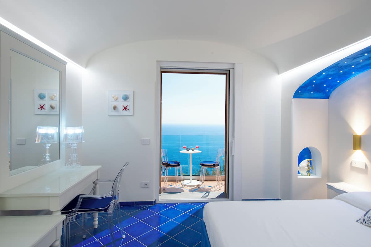 Une chambre d’hôtel moderne avec un balcon, un grand lit, une salle de bain avec un miroir et un lavabo, et une vue sur la mer.