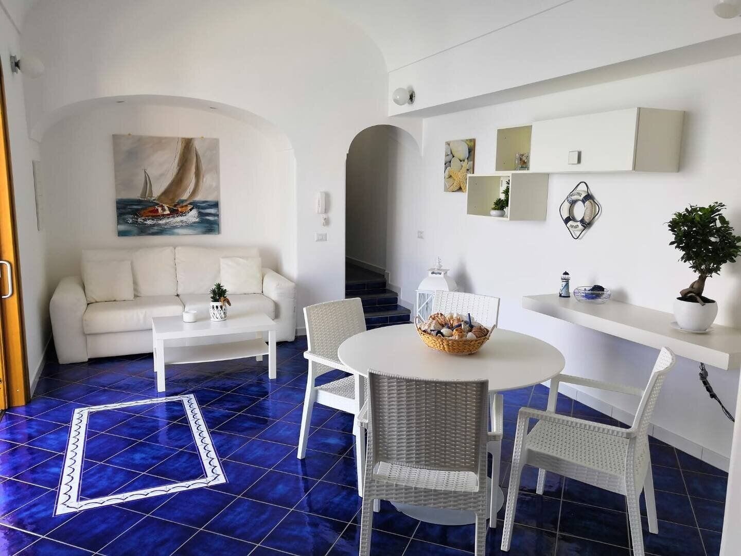 Ein modernes Wohnzimmer mit einem weißen Sofa, einem runden Esstisch mit Korbstühlen und einem blauen Fliesenboden.