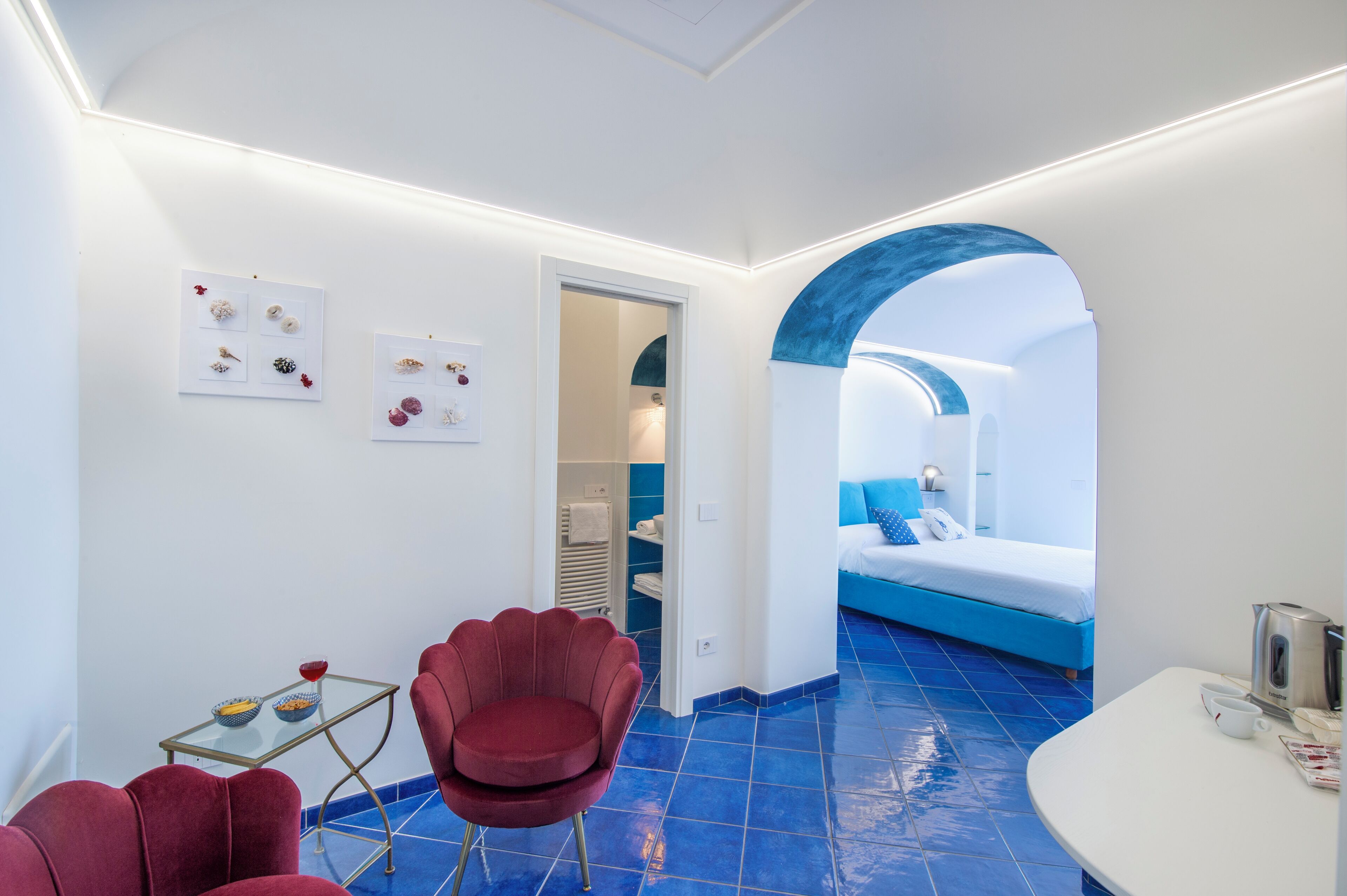 Une chambre d’hôtel moderne avec un sol carrelé bleu, un fauteuil rouge, une table en verre avec un bol et une tasse, et un lit avec une literie bleue.