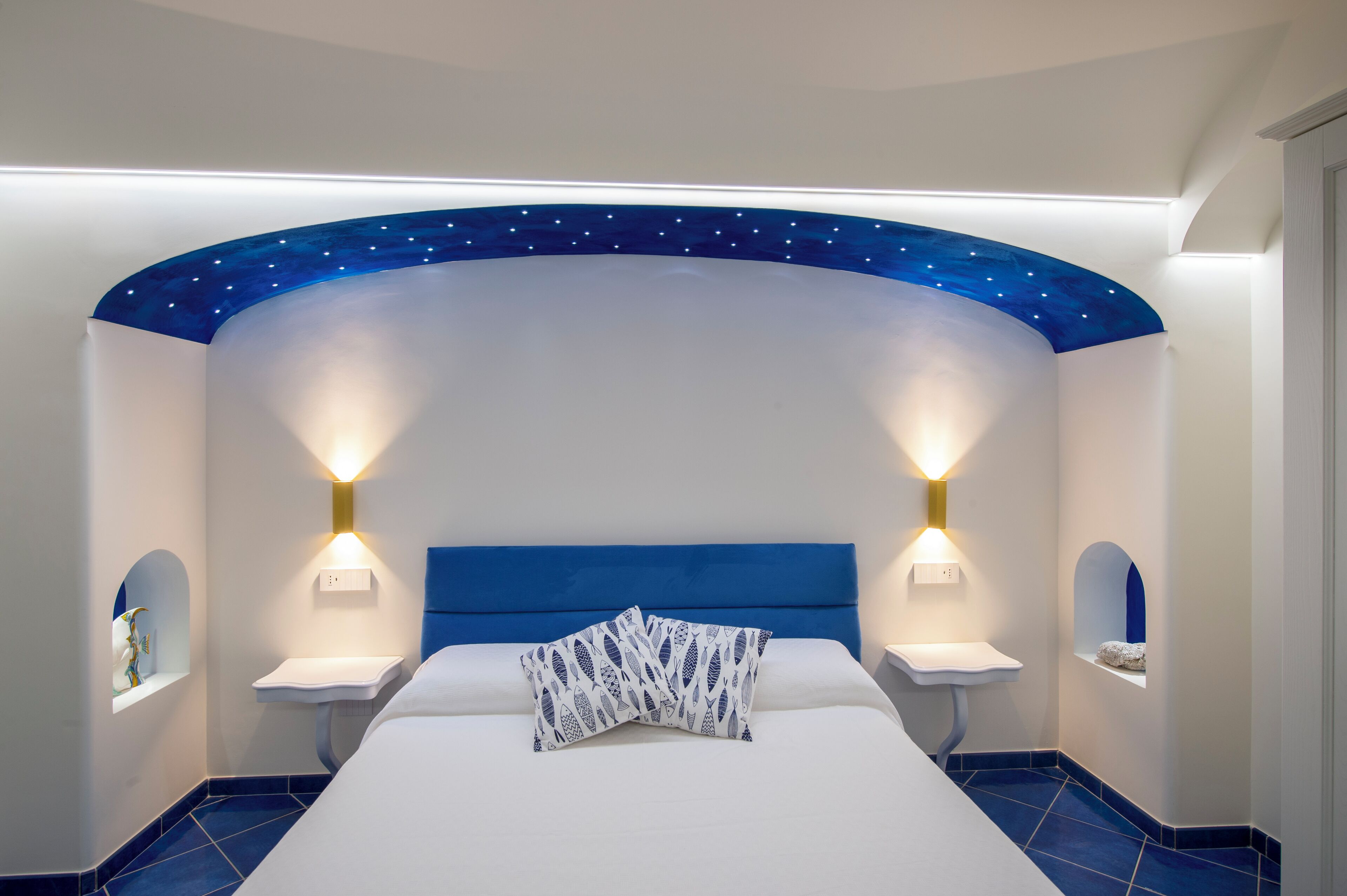 Une chambre à coucher avec un plafond incurvé, deux lampes de chevet, un lit avec une tête de lit blanche et un sol carrelé de couleur bleue.