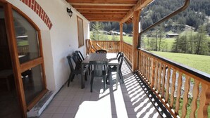 A covered outdoor patio with a wooden table and chairs, a view of a green landscape, and a mountain in the background. - Gites Simond welcomes you to Arvieux in the heart of the Queyras Regional Park, (ARVIEUX)